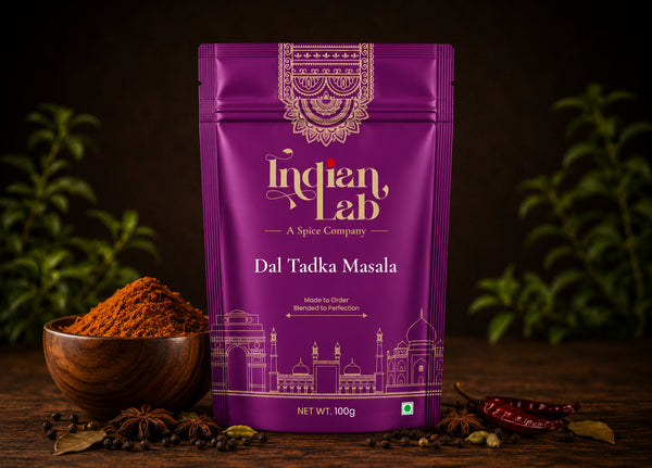 A purple stand-up pouch of Indian Lab Dal Tadka Masala spice mix stands on a dark wooden surface, with a small wooden bowl of orange-brown spice powder, star anise, and red chilies arranged around it. The pouch has ornate gold and white branding and a line drawing of the Taj Mahal at the bottom.