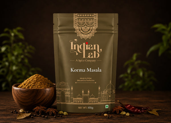 A 100g stand-up pouch of Indian Lab Korma Masala spice mix displayed on a rustic wooden table. The pouch is olive green with gold illustrations of Indian architecture and the brand name 'Indian Lab'.