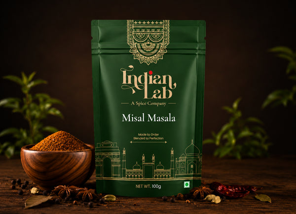 A 100g pouch of Indian Lab Misal Masala spice mix, displayed on a rustic wooden table with a wooden bowl of loose powder, whole spices, and green leaves against a dark background.