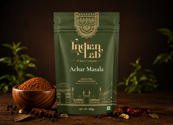 A 100g pouch of Indian Lab Achar Masala spice mix, displayed on a wooden surface with a wooden bowl of the ground spice, whole spices, and red chilies, set against a dark background with blurred green plants.