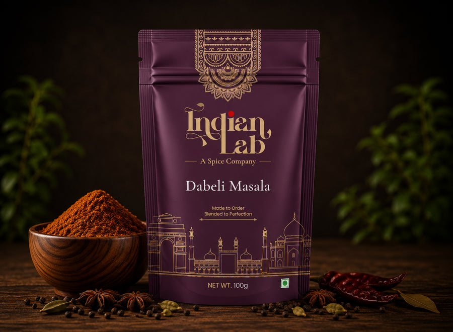 A 100g stand-up pouch of Indian Lab Dabeli Masala spice mix, displayed on a rustic wooden table with a wooden bowl of ground spice, whole spices, and red chilies against a dark, blurred background.