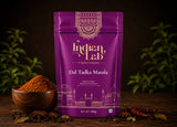 A purple stand-up pouch of Indian Lab Dal Tadka Masala spice mix stands on a dark wooden surface, with a small wooden bowl of orange-brown spice powder, star anise, and red chilies arranged around it. The pouch has ornate gold and white branding and a line drawing of the Taj Mahal at the bottom.