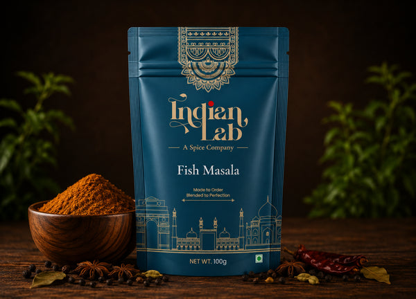 A 100g stand-up pouch of Indian Lab Fish Masala spice mix, displayed on a rustic wooden table with a wooden bowl of orange-brown powder, whole spices, and dried red chilies against a dark background with soft green foliage.