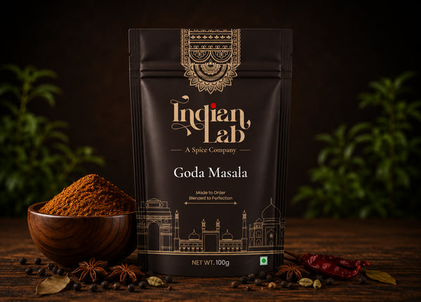 A 100g stand-up pouch of Indian Lab Goda Masala spice mix, styled on a dark wooden table with a wooden bowl of powder, whole spices, and dried red chilies against a dark background with green foliage.