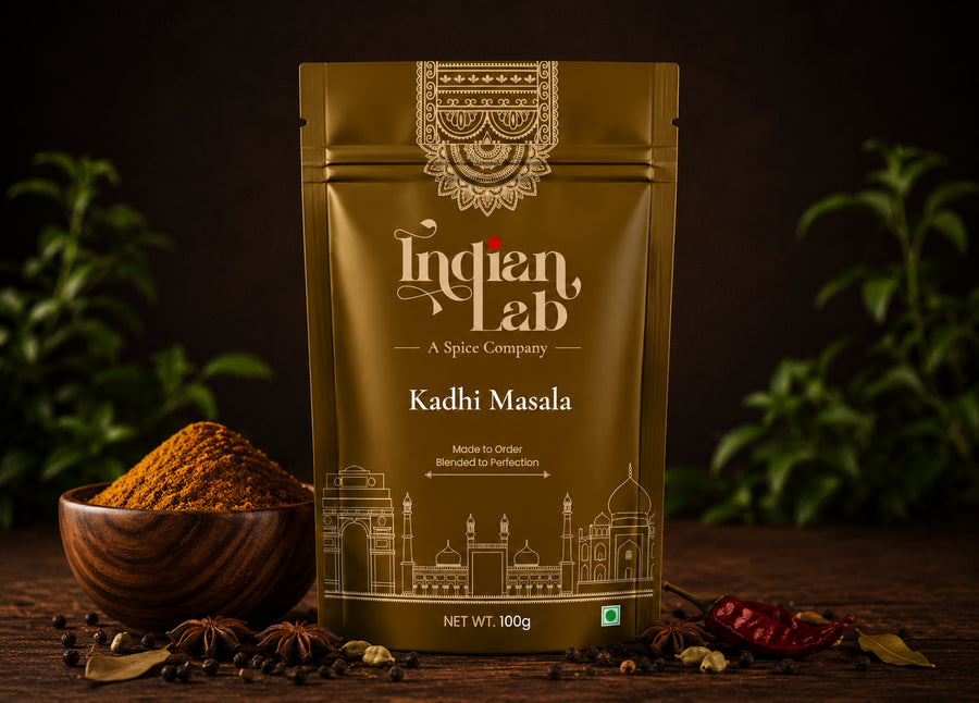 A 100g stand-up pouch of Indian Lab Kadhi Masala spice mix, displayed on a dark wooden surface with a wooden bowl of orange-brown powder, whole spices, and red chilies, set against a dark background with soft green foliage.
