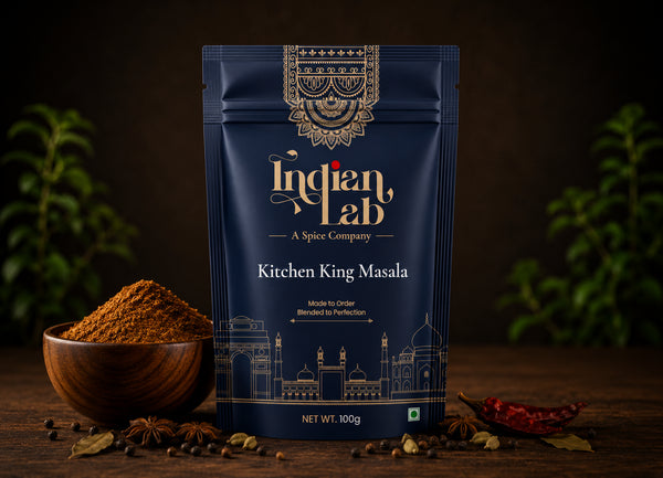A navy blue stand-up pouch of Indian Lab Kitchen King Masala spice mix, styled on a dark wooden surface with a bowl of the brown spice powder, star anise, and dried red chilies, with blurred green plants in the background.