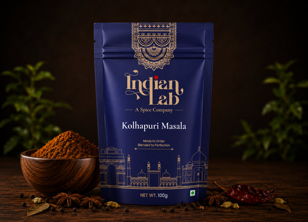 A 100g stand-up pouch of Indian Lab Kolhapuri Masala spice mix, displayed on a rustic wooden table with a wooden bowl of the ground spice, star anise, and red chili peppers. The background is dark with soft green foliage bokeh.