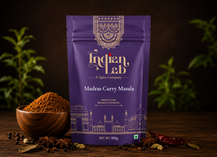 A purple stand-up pouch of Indian Lab Madras Curry Masala spice mix, styled on a dark wooden surface with a wooden bowl of the powder, whole spices, and dried red chilies, with blurred green plants in the background.