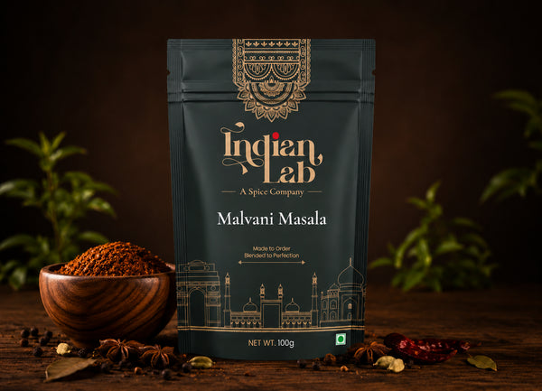 A 100g pouch of Indian Lab Malvani Masala spice mix, styled on a dark wooden table with a wooden bowl of the ground spice, whole spices, and green leaves in the background.