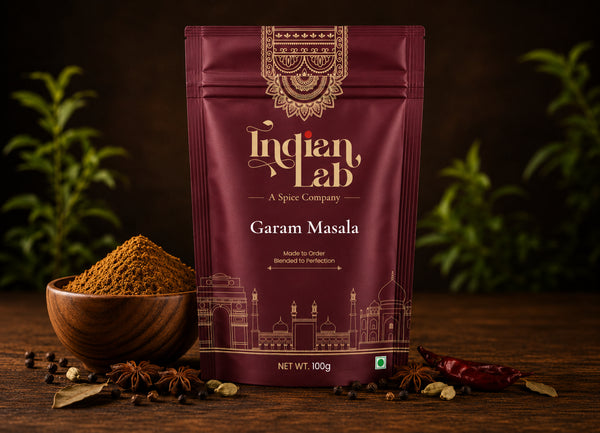 A 100g stand-up pouch of Indian Lab Garam Masala spice mix on a dark wooden table, styled with a wooden bowl of the ground spice, star anise, and dried red chilies, with green plants blurred in the background.