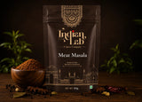 A 100g stand-up pouch of Indian Lab Meat Masala spice mix, displayed on a dark wooden table with a wooden bowl of loose powder, whole spices, and green leaves in the background.