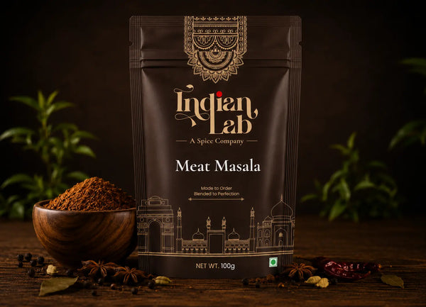 A 100g stand-up pouch of Indian Lab Meat Masala spice mix, displayed on a dark wooden table with a wooden bowl of loose powder, whole spices, and green leaves in the background.