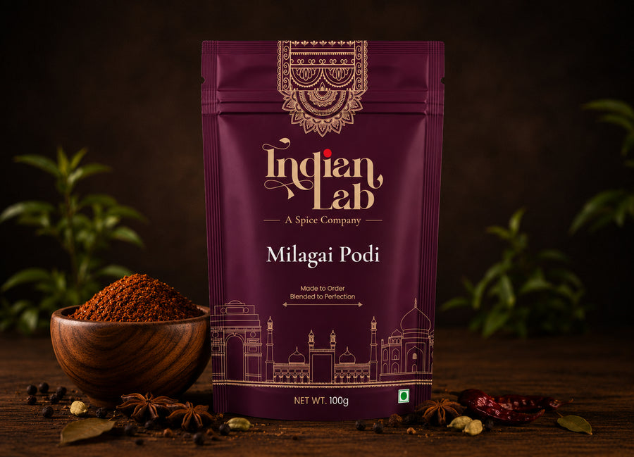 A 100g stand-up pouch of Indian Lab Milagai Podi spice mix, displayed on a rustic wooden surface with a wooden bowl of the spice, star anise, and whole spices, set against a dark background with blurred green plants.
