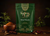 A 100g pouch of Indian Lab Misal Masala spice mix, displayed on a rustic wooden table with a wooden bowl of loose powder, whole spices, and green leaves against a dark background.