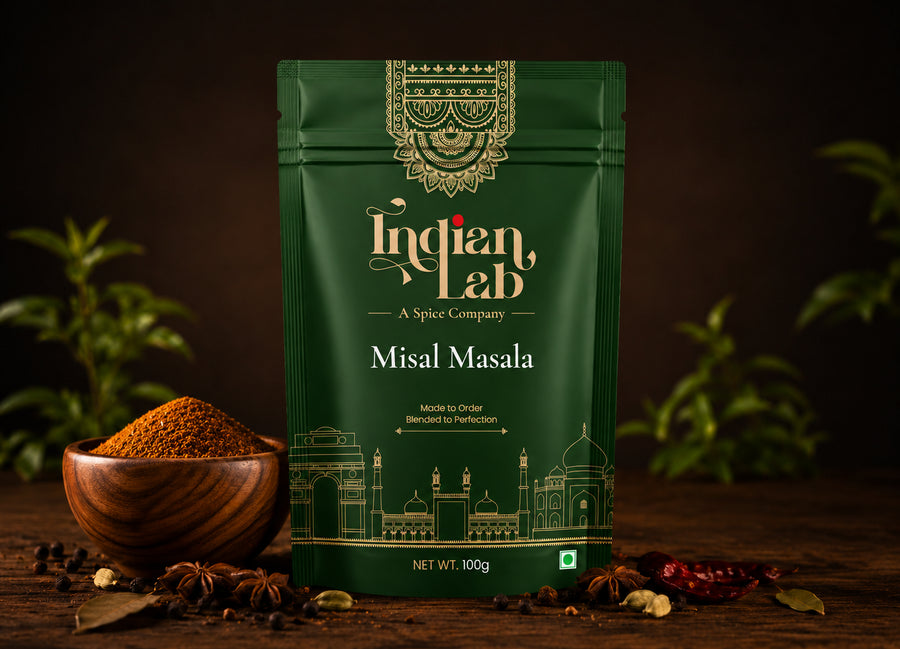 A 100g pouch of Indian Lab Misal Masala spice mix, displayed on a rustic wooden table with a wooden bowl of loose powder, whole spices, and green leaves against a dark background.