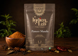 A stand-up pouch of Indian Lab Paneer Masala spice mix on a dark wooden surface, surrounded by a small wooden bowl of orange-brown powder, whole spices, and dried red chilies, with blurred green plants in the background.