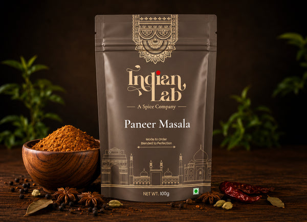 A stand-up pouch of Indian Lab Paneer Masala spice mix on a dark wooden surface, surrounded by a small wooden bowl of orange-brown powder, whole spices, and dried red chilies, with blurred green plants in the background.