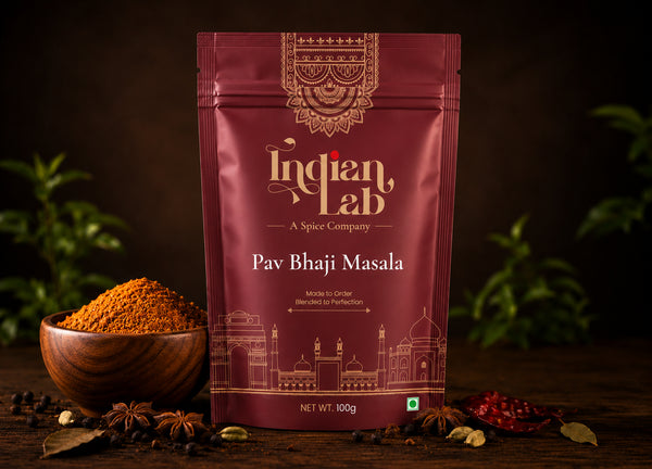 A 100g pouch of Indian Lab Pav Bhaji Masala spice mix stands on a dark wooden table, flanked by a wooden bowl filled with the orange-brown masala powder, whole spices, and dried red chilies. The background is dark with soft green foliage bokeh.