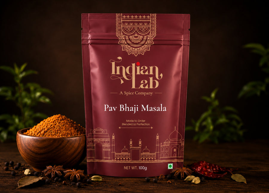 A 100g pouch of Indian Lab Pav Bhaji Masala spice mix stands on a dark wooden table, flanked by a wooden bowl filled with the orange-brown masala powder, whole spices, and dried red chilies. The background is dark with soft green foliage bokeh.