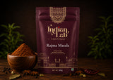 A 100g stand-up pouch of Indian Lab Rajma Masala spice mix displayed on a rustic wooden table, flanked by a wooden bowl filled with the ground red-brown spice powder, whole peppercorns, star anise, and dried red chilies, with a dark, moody background and blurred green herbs.