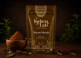A 100g pouch of Indian Lab Rasam Masala spice mix stands on a dark wooden surface, flanked by a wooden bowl filled with the brown powder, whole spices, and dried red chilies, with blurred green herbs in the background.