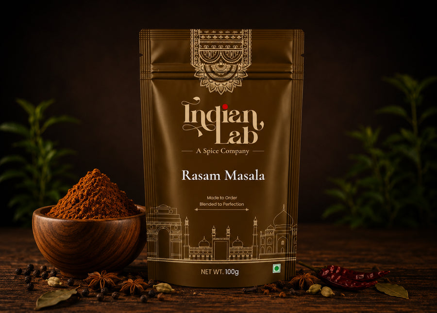 A 100g pouch of Indian Lab Rasam Masala spice mix stands on a dark wooden surface, flanked by a wooden bowl filled with the brown powder, whole spices, and dried red chilies, with blurred green herbs in the background.