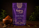 A purple stand-up pouch of Indian Lab Sambar Masala spice mix stands on a dark wooden surface, with a small wooden bowl of reddish-brown powder, peppercorns, and whole spices around it. The pouch has ornate gold detailing and the brand name 'Indian Lab' is clearly visible.