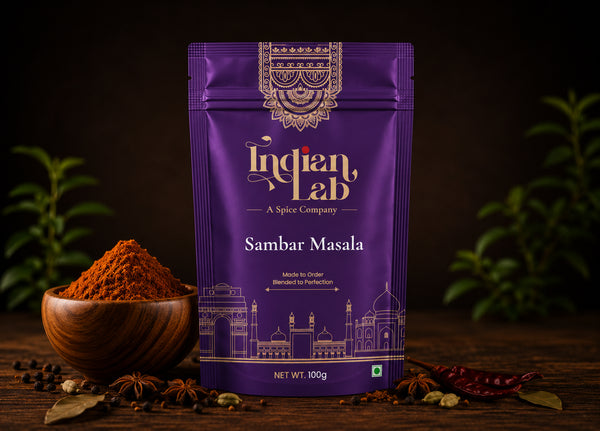 A purple stand-up pouch of Indian Lab Sambar Masala spice mix stands on a dark wooden surface, with a small wooden bowl of reddish-brown powder, peppercorns, and whole spices around it. The pouch has ornate gold detailing and the brand name 'Indian Lab' is clearly visible.