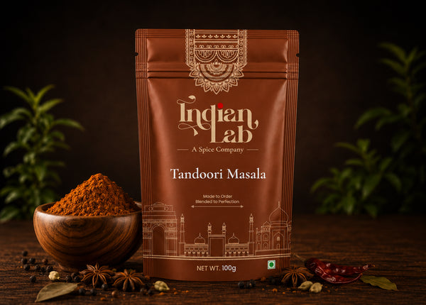 A 100g pouch of Indian Lab Tandoori Masala spice mix, displayed on a dark wooden table with a wooden bowl of the powder, star anise, and dried red chilies. The pouch is brown with gold and white text and graphics, set against a dark background with blurred green plants.