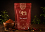 A 100g pouch of Indian Lab Tikka Masala spice mix on a dark wooden table, styled with a wooden bowl of powder, star anise, and dried red chilies, with green plants blurred in the background.