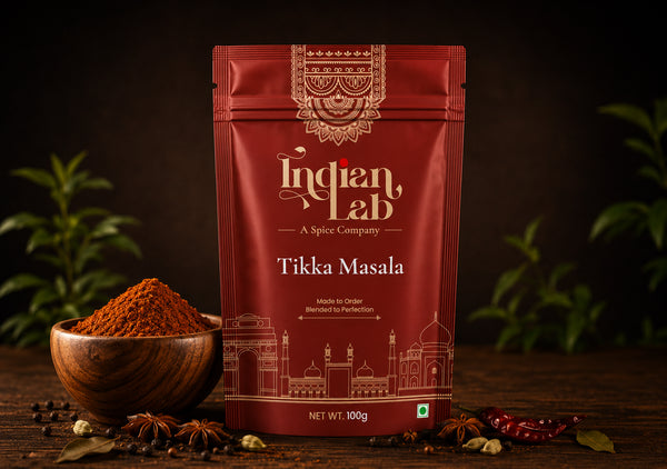 A 100g pouch of Indian Lab Tikka Masala spice mix on a dark wooden table, styled with a wooden bowl of powder, star anise, and dried red chilies, with green plants blurred in the background.