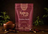 A 100g stand-up pouch of Indian Lab Vindaloo Masala spice mix, displayed on a dark wooden surface with a wooden bowl of the spice, star anise, and dried red chilies, set against a dark background with blurred green plants.