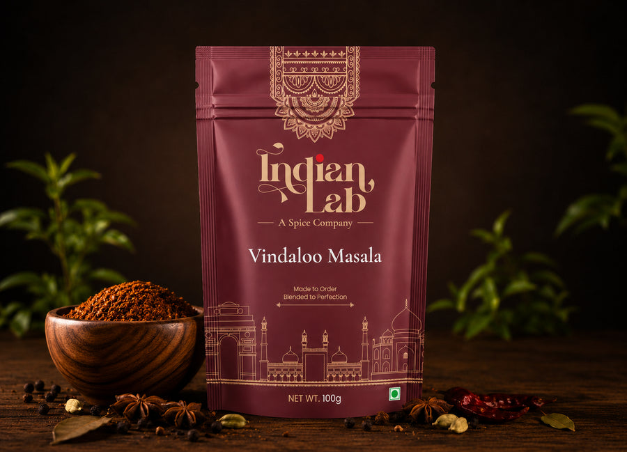 A 100g stand-up pouch of Indian Lab Vindaloo Masala spice mix, displayed on a dark wooden surface with a wooden bowl of the spice, star anise, and dried red chilies, set against a dark background with blurred green plants.