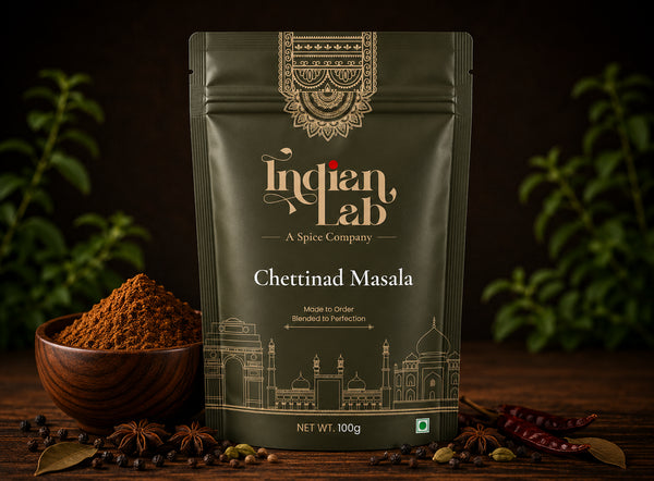 A 100g stand-up pouch of Indian Lab Chettinad Masala spice mix, styled on a dark wooden table with a bowl of loose powder, whole spices, and dried red chilies against a dark background with soft green foliage.
