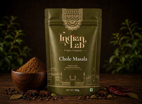 A 100g stand-up pouch of Indian Lab Chole Masala spice mix, styled on a dark wooden surface with a small bowl of the brown powder, whole spices, and dried red chilies against a dark background with green foliage.