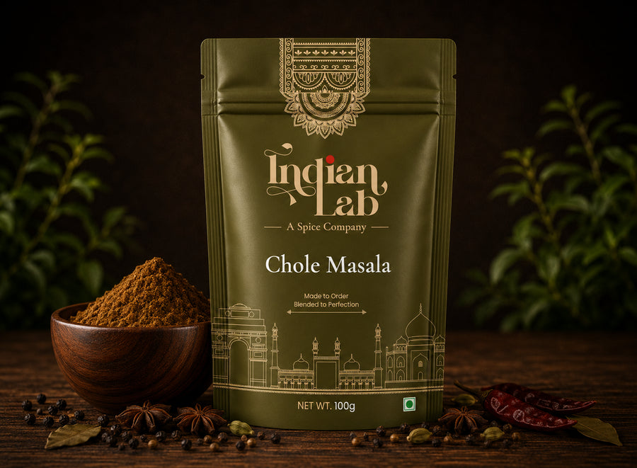A 100g stand-up pouch of Indian Lab Chole Masala spice mix, styled on a dark wooden surface with a small bowl of the brown powder, whole spices, and dried red chilies against a dark background with green foliage.