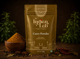 A 100g stand-up pouch of Indian Lab Curry Powder, styled on a dark wooden table with a wooden bowl of curry powder, whole spices, and dried red chilies against a dark, blurred background with green plants.